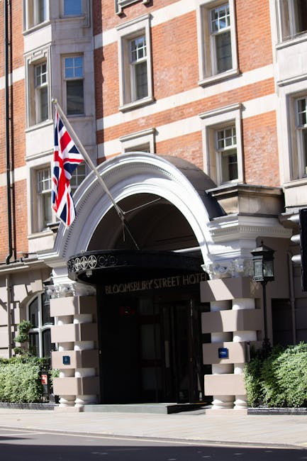 The entrance of the Brunswick Street Hotel in Bloomsbury features a white archway canopy with decorative molding, situated above a dark doorway. To the left side of the arch, a Union Jack flag is mounted on a flagpole. The building's facade includes multiple brick and stonework elements with several windows visible on the upper floors. In front of the entrance, there are small green shrubs on either side, and the sidewalk is clear. This scene captures the exterior of a hotel in a city environment, relevant to house removals and relocation services, with the hotel’s entryway prepared for client or guest access, supporting logistics and home relocation planning.