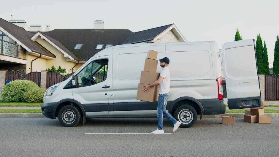 A man wearing a white T-shirt, blue jeans, and a black cap is outside a white cargo van parked on a residential street, carrying three large cardboard boxes stacked vertically. The van, which belongs 