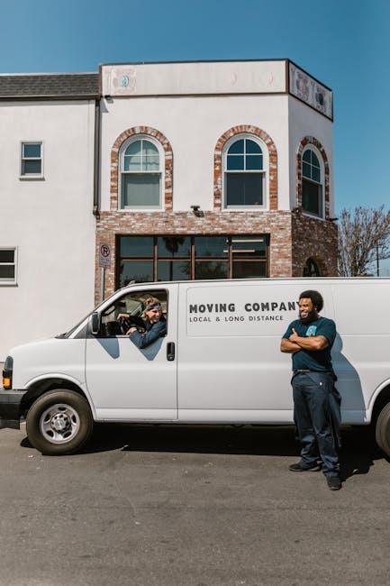 A man wearing a white T-shirt, blue jeans, and a black cap is outside a white cargo van parked on a residential street, carrying three large cardboard boxes stacked vertically. The van, which belongs 