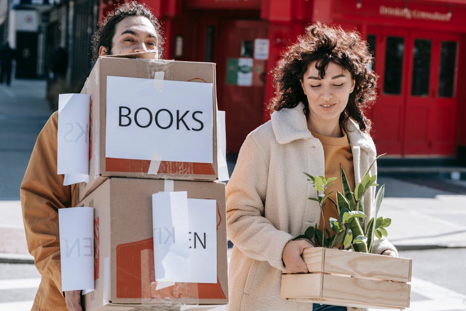 Two individuals participating in a house relocation process outdoors on a city street, with a red storefront in the background. The person on the left, partially obscured, is holding a stack of cardboard moving boxes secured with packing tape and labeled with white paper signs reading 'BOOKS' and 'KIM.' These boxes are stacked one on top of the other, and the individual is supporting them with both hands. The person on the right, a woman with curly brown hair, is holding a wooden plant pot containing green leafy plants, dressed in a beige coat over a yellow top. The scene captures the loading process as part of furniture transport and packing during a home move, with natural daylight illuminating the area. This image illustrates the careful handling of household items, including books and indoor plants, during professional removals, with the involvement of a company like Man With a Van Bloomsbury, dedicated to house removals and moving services.