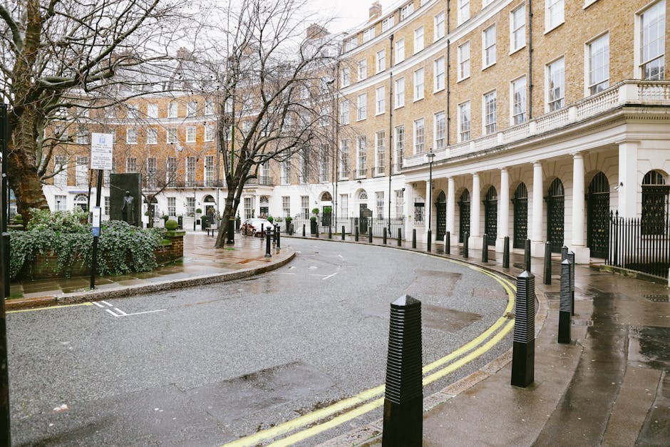 A wet, empty street scene in Bloomsbury, London, focusing on the curved roadway bordered by black bollards along the pavement, which is slick from recent rain. The street is lined with a white, multi-storey Georgian-style building featuring large windows and a row of white columns supporting a covered walkway. Nearby, there is a small landscaped area with bushes and a leafless tree, along with a signpost indicating parking restrictions. In the background, more buildings with similar architectural styles are visible, with several parked cars and a few pedestrians under umbrellas. The environment suggests a calm moment during a home relocation process, where furniture or moving boxes might be loaded into a nearby van for transport. Man With a Van Bloomsbury's removal services are relevant to this setting, which highlights the logistics of packing and moving in an urban area with regulated street access.