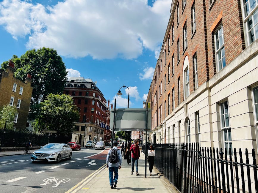 Two individuals participating in a house relocation process outdoors on a city street, with a red storefront in the background. The person on the left, partially obscured, is holding a stack of cardboard moving boxes secured with packing tape and labeled with white paper signs reading 'BOOKS' and 'KIM.' These boxes are stacked one on top of the other, and the individual is supporting them with both hands. The person on the right, a woman with curly brown hair, is holding a wooden plant pot containing green leafy plants, dressed in a beige coat over a yellow top. The scene captures the loading process as part of furniture transport and packing during a home move, with natural daylight illuminating the area. This image illustrates the careful handling of household items, including books and indoor plants, during professional removals, with the involvement of a company like Man With a Van Bloomsbury, dedicated to house removals and moving services.