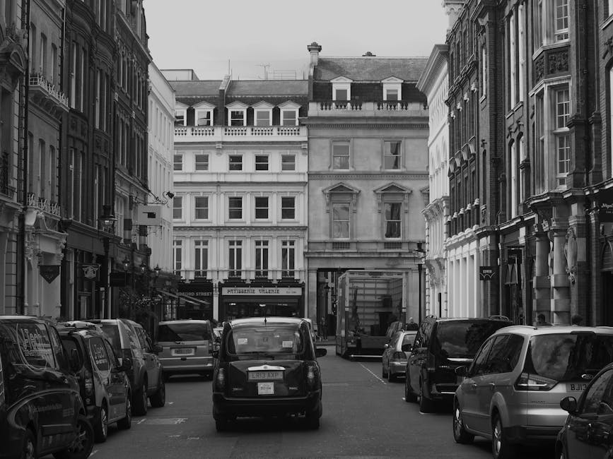 Black and white photograph of a narrow urban street with tightly parked cars lining both sides, including small vans and passenger vehicles. Behind the parked vehicles, there are multi-storey period-s