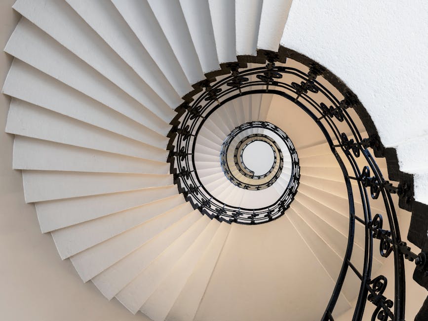 View looking directly down a multi-story stairwell inside a building, showing a square-shaped spiral staircase with black metal railings and wooden handrails. The staircase's steps are painted beige, 